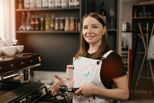 Um barista com avental branco sorri e posa segurando uma calcadeira de metal e um porta-filtro com café