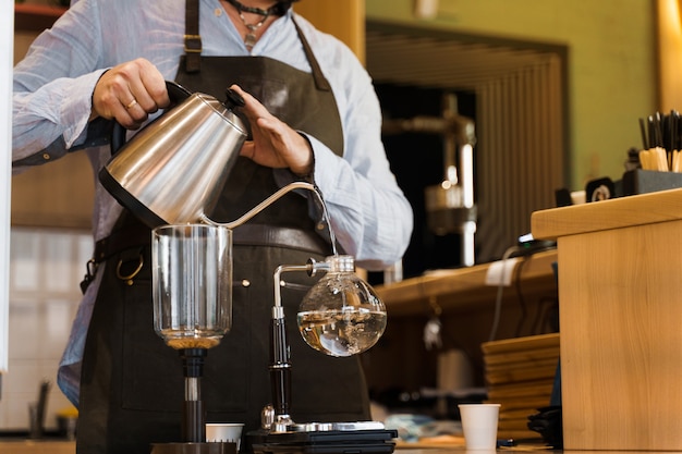 Um barista de close-up derrama água fervente de kattle em um dispositivo de sifão de vidro para fazer café em uma cafeteria