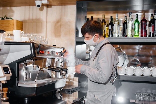 Um barista mascarado prepara um delicioso café no bar de uma cafeteria. O trabalho de restaurantes e cafés durante a pandemia.