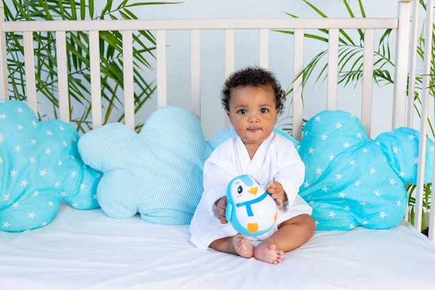 Um bebê africano em uma túnica branca após um banho e banho senta-se na cama em casa antes de ir para a cama e brinca com um brinquedo um conceito de cuidado e higiene para crianças pequenas