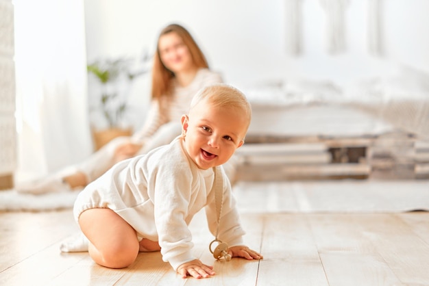 Um bebê alegre rasteja no chão e ri da câmera com a mãe ao fundo