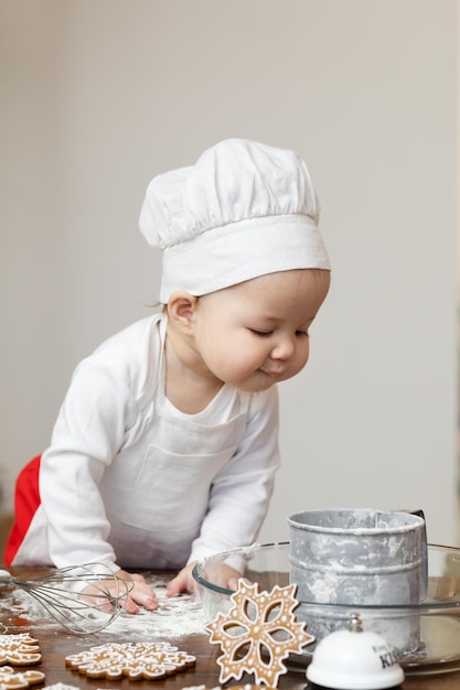 Um bebê asiático com chapéu e avental de chef está preparando biscoitos de gengibre de Natal Com curiosidade, ele olha para o recipiente com farinha