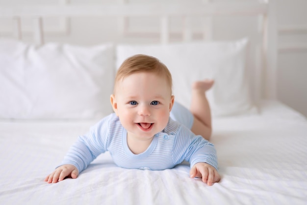 Um bebê bonito e saudável está deitado em uma cama na cama branca em casa em uma roupa azul O garoto olha para a câmera sorri