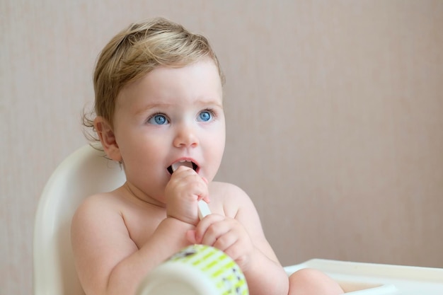 Um bebê com olhos azuis e cabelos loiros se senta em uma cadeira alta e segura uma colher na boca