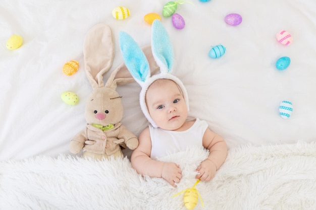 Um bebê com orelhas de coelho na cabeça está deitado em um berço com um brinquedo de coelho e ovos de páscoa um bebê engraçado e fofo conceito de páscoa