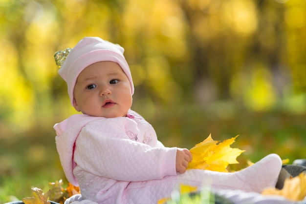 Um bebê com roupas rosa sentado entre o outono amarelo, folhas caem em uma cena do parque.