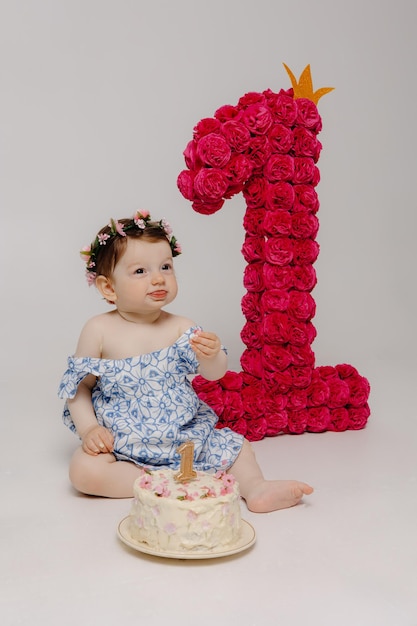 Foto um bebê com uma flor no cabelo e o número 1 está sentado na frente de um bolo