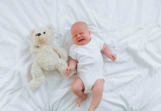 Um bebê de 2 meses está chorando na cama com uma vista superior de ursinho de pelúcia