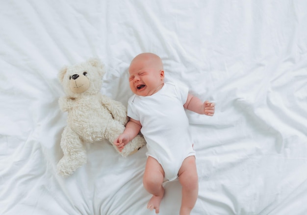 Um bebê de 2 meses está chorando na cama com uma vista superior de ursinho de pelúcia