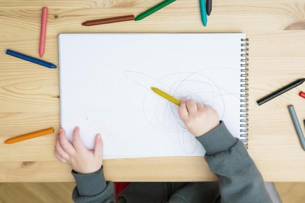 Um bebê desenha com giz de cera colorido em um álbum sobre uma mesa de madeira Vista superior