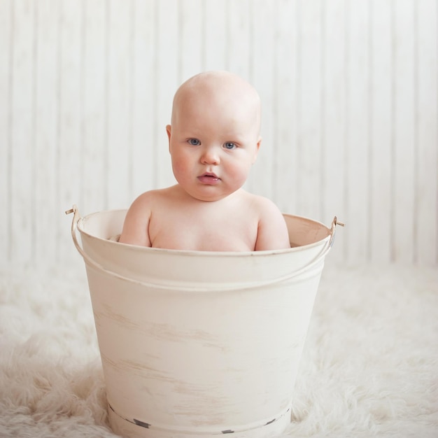 Um bebê em um balde com olhos azuis está sentado em um tapete branco.