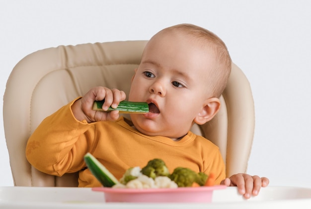 Um bebê em uma cadeira de alimentação come um pepino fresco