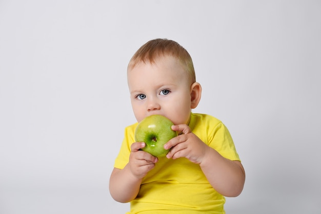 Um bebê em uma camiseta amarela segura uma maçã verde em suas mãos A criança está sentada em um fundo branco com uma maçã verde