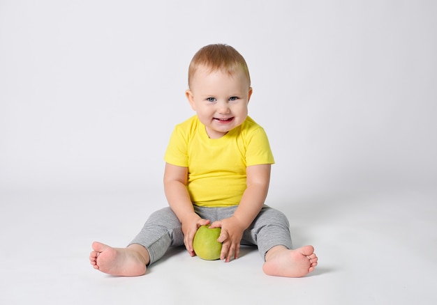 Um bebê em uma camiseta amarela segura uma maçã verde em suas mãos A criança está sentada em um fundo branco com uma maçã verde