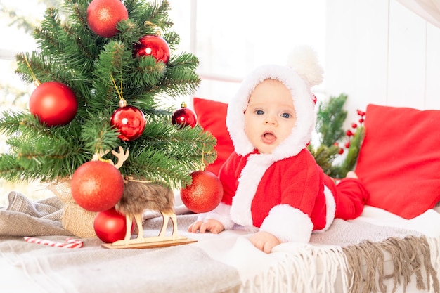 Um bebê em uma fantasia de Papai Noel debaixo da árvore de Natal brinca com bolas vermelhas de Natal na janela da casa o conceito do ano novo