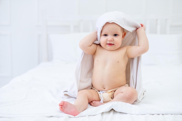 Um bebê em uma toalha branca depois de tomar banho em uma banheira em uma cama brilhante em casa sorri o conceito de higiene e lavagem