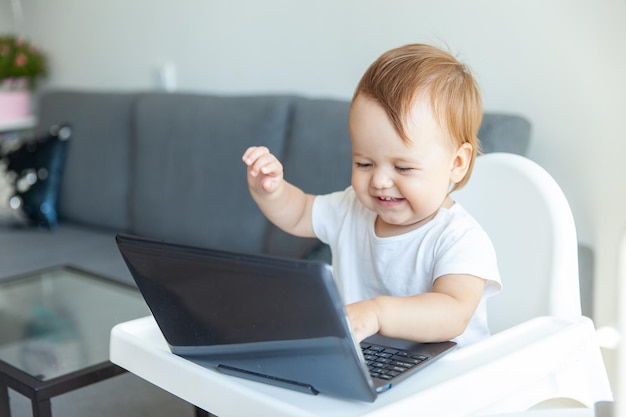 Um bebê engraçado usa um laptop enquanto está sentado na mesa das crianças em casa