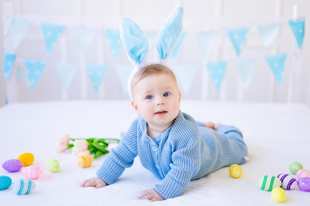 Um bebê feliz com orelhas de coelhinho da páscoa ovos de páscoa coloridos está em casa em uma cama em linho branco pastel cartão de páscoa