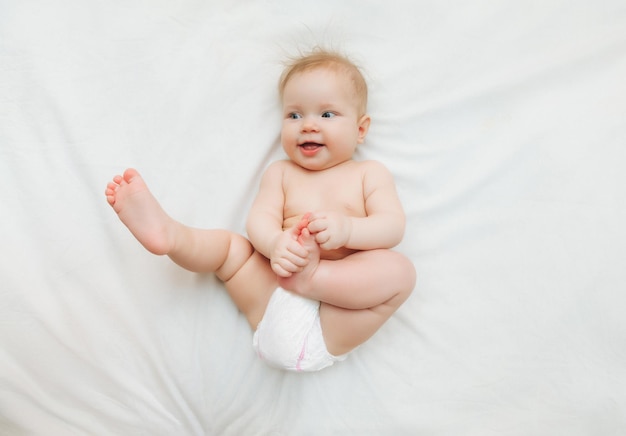 Um bebê feliz e alegre em fraldas está deitado em uma cama branca e segurando sua perna Lugar de foto de alta qualidade para texto