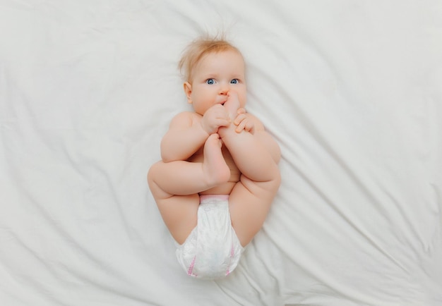 Um bebê feliz em fraldas está deitado em uma cama branca e segurando sua perna Lugar de foto de alta qualidade para texto