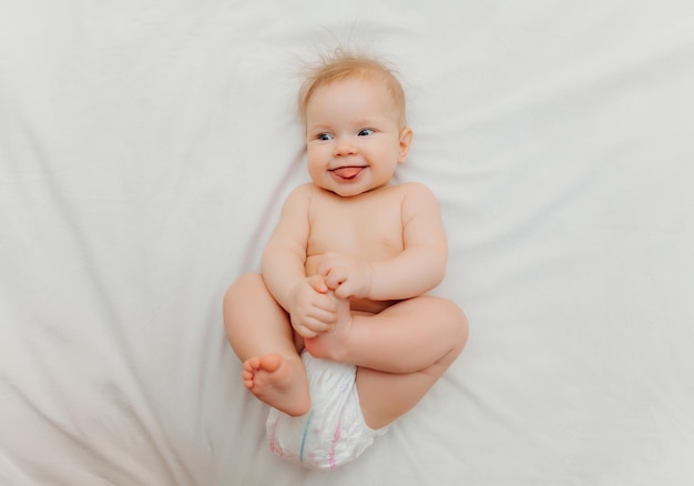 Um bebê feliz em fraldas está deitado em uma cama branca e segurando sua perna Lugar de foto de alta qualidade para texto