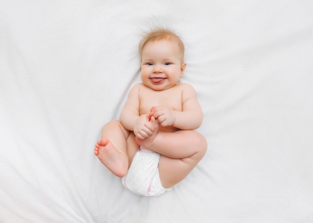 Um bebê feliz em fraldas está deitado em uma cama branca e segurando sua perna Lugar de foto de alta qualidade para texto