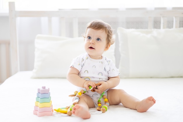 Um bebê fofo e saudável está sentado na cama, na roupa de cama, sorrindo, brincando com brinquedos para crianças