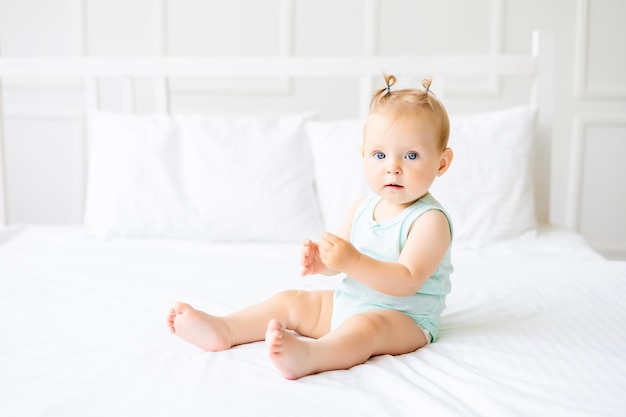 Um bebê fofo em um body feito de tecido de algodão natural está sentado na roupa de cama branca no quarto Um bebê feliz na cama Têxteis e roupa de cama para crianças