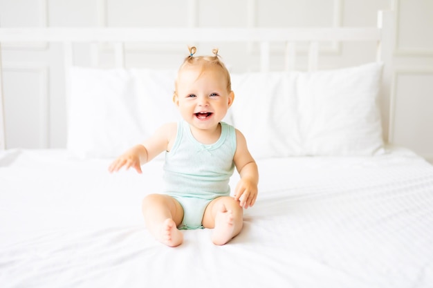 Um bebê fofo em um body feito de tecido de algodão natural está sentado na roupa de cama branca no quarto Um bebê feliz na cama Têxteis e roupa de cama para crianças