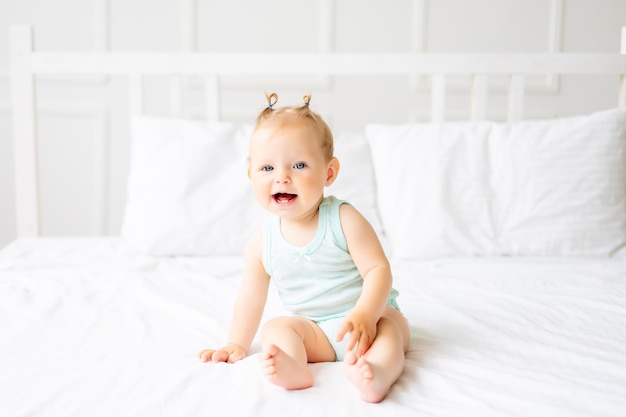 Um bebê fofo em um body feito de tecido de algodão natural está sentado na roupa de cama branca no quarto Um bebê feliz na cama Têxteis e roupa de cama para crianças