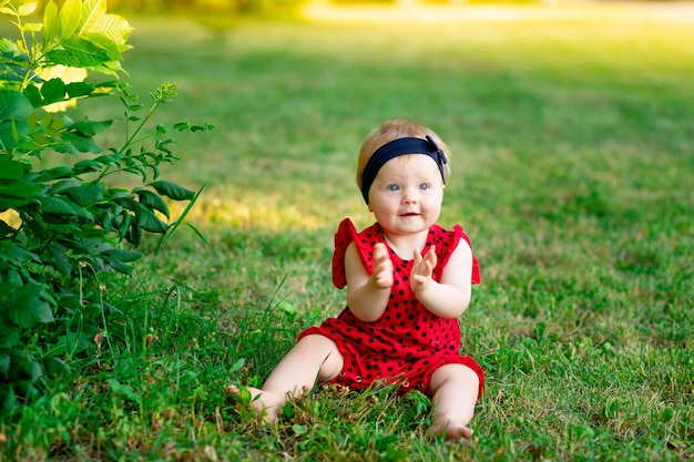 Um bebê no verão na grama verde em uma roupa vermelha bate palmas de alegria no espaço do sol poente para texto