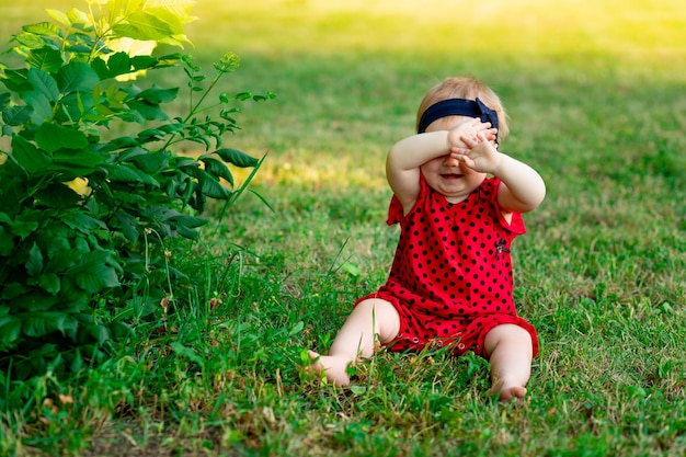 Um bebê no verão na grama verde em uma roupa vermelha cobriu o rosto com as mãos escondidas no sol poente