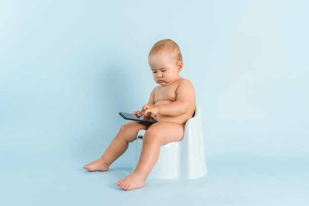 Um bebê recém-nascido com um telefone está sentado em um penico em um fundo azul. Uma criança de 12 a 15 meses está brincando em um smartphone enquanto está sentada em um penico