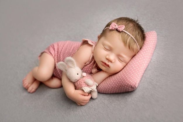 Um bebê recém-nascido fofo em um terno rosa e uma faixa rosa com uma borboleta na cabeça dorme docemente Coelho de pera de malha nas mãos Foto macro profissional em um fundo cinza