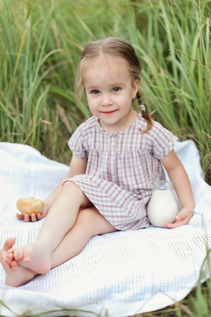 Um bebê tem uma jarra de leite nas mãos em um dia de verão em um campo verde. Uma menina está sentada em um cobertor e curtindo a natureza