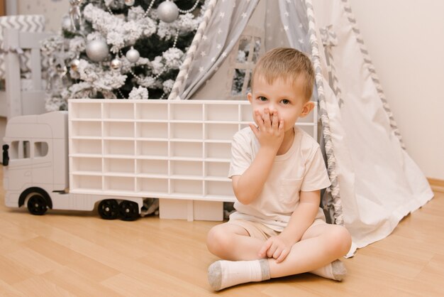 Um bebezinho fofo senta-se no quarto das crianças em uma tenda e manda um beijo ao lado de uma árvore de neve de Natal e um carro de madeira