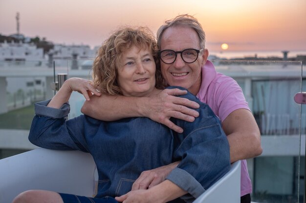 Um belo casal de idosos caucasianos abraçados no terraço da casa a desfrutar do pôr-do-sol.