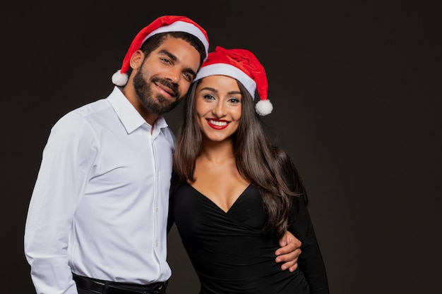 Um belo casal de jovens hispânicos com chapéus de Papai Noel a abraçar-se em preto.