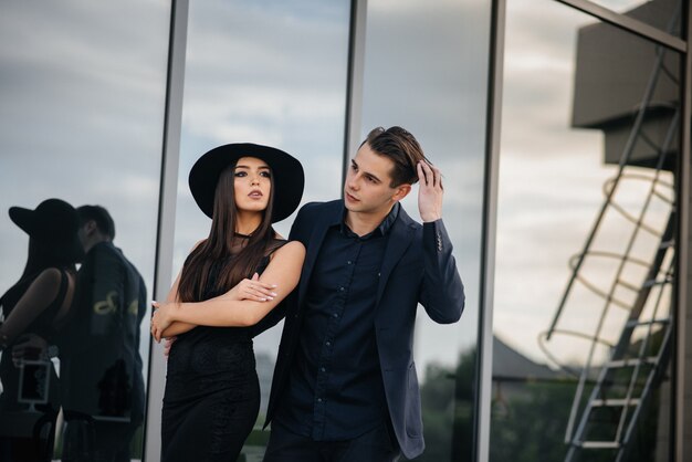 Um belo e elegante par de jovens em roupas e óculos pretos fica no contexto de um prédio de escritórios por do sol. moda e estilo