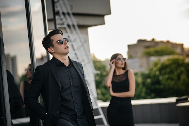 Um belo e elegante par de jovens em roupas pretas e óculos fica contra um prédio de escritórios por do sol. moda e estilo