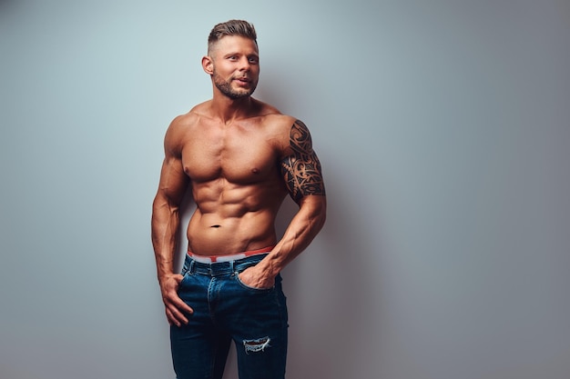 Um belo fisiculturista sem camisa com corte de cabelo elegante e barba, com tatuagem no braço, posando em um estúdio. Isolado em um fundo cinza.