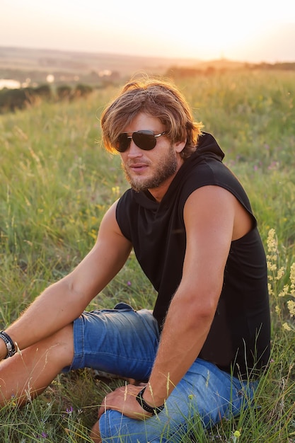 um belo jovem loiro de óculos e uma camiseta preta está sentado na grama ao pôr do sol