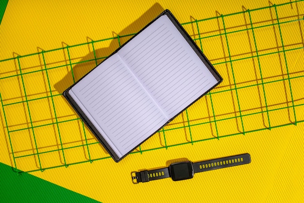 Um bloco de notas aberto e um relógio inteligente em uma grade de metal em um fundo amarelo