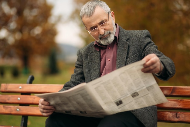 Um bom avô com uma bela barba em uma jaqueta cinza senta-se em um banco no parque e lê um