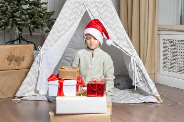 Um bonito menino pré-escolar com um chapéu de papai noel sentado no chão ao lado de uma pilha de seus presentes no berçário a criança está feliz com os presentes do conceito de natal do papai noel kids