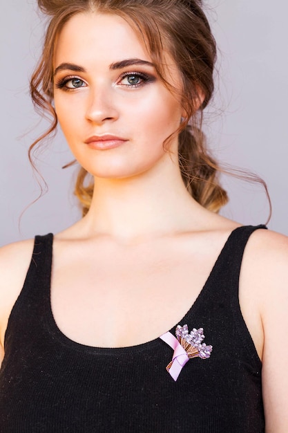 Um broche na forma de um buquê de flores lilás em uma camiseta preta de uma menina morena A menina está posando em um fundo cinza sorrindo e olhando para a câmera