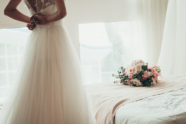 Um buquê de casamento em uma cama, com noiva turva, abotoando o vestido, vista traseira