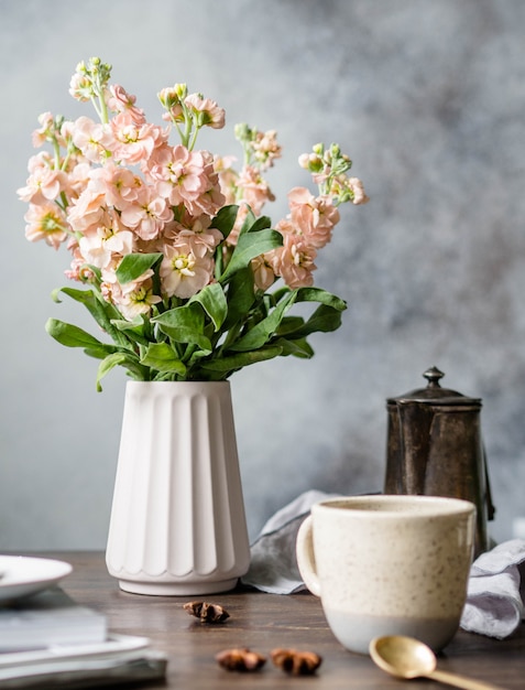 Um buquê de flores mattiol rosa em um vaso, um pote de café vintage, copo com café e especiarias em uma mesa de madeira marrom.