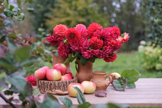 Um buquê de flores vermelhas em um jarro e uma cesta com maçãs em uma mesa de madeira no jardim