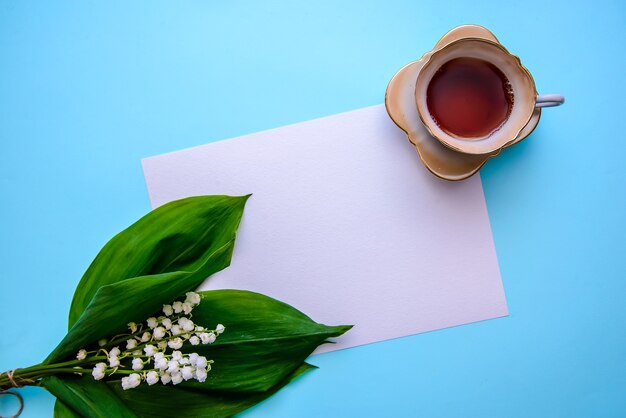 Um buquê de lindos lírios do vale com folhas verdes, uma xícara de chá e uma folha de papel branca sobre uma superfície azul